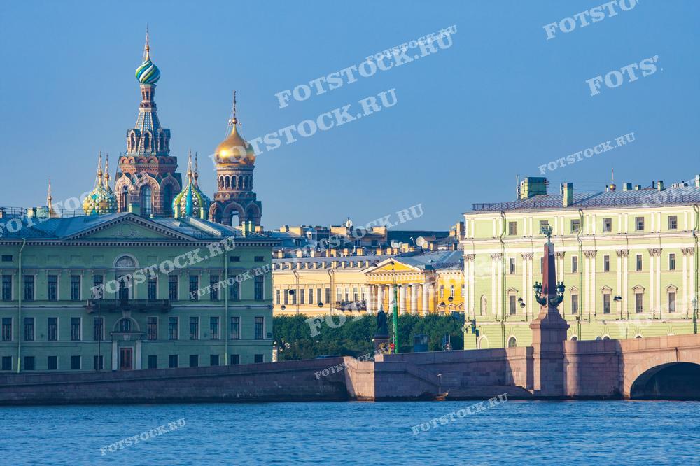 На фотографии изображен храм Спас-на-Крови и набережная реки Невы в Санкт-Петербурге. Летний день подчеркивает красоту разноцветных куполов храма и окружающих зданий. На переднем плане виден один из мостов города, соединяющий берега Невы. Яркое солнце освещает архитектурные шедевры Петербурга.